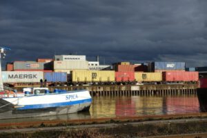 Das Bild zeigt Container, die im Niehler Hafen stehen sowie ein Boot im Vordergrund auf dem Rhein.