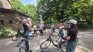 Das Bild zeigt die Exkursionsgruppe am Fort in Köln-Nippes neben ihren Fahrrädern stehen. Winni, der Exkursionsleiter, hat die Blicke auf sich gerichtet und gestikuliert herum, während er Geschichten erzählt.
